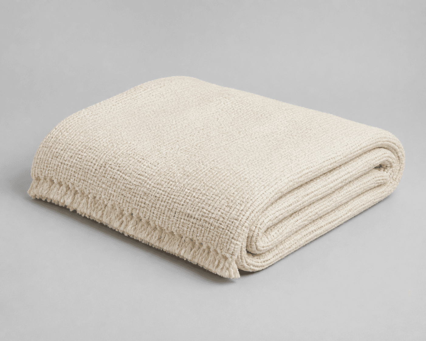 Folded light-neutral textured throw blanket showing a woven surface, rounded rolled fold, and finished fringe edge on a quiet neutral background